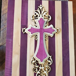 Puede incluir: Una cruz de madera decorativa con un diseño en capas, con un centro magenta vibrante y un borde de madera clara. La cruz se encuentra sobre un fondo a rayas de tonos púrpuras y madera natural alternados, creando un contraste llamativo. Estilo ornamental.
