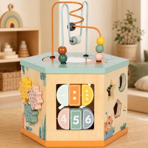 May include: A hexagonal wooden activity cube featuring a bead maze on top with orange and blue wires. The sides include rotating gears, a number matching puzzle with digits 4, 5, and 6, and various shape cutouts. The natural wood finish is accented with muted tones of sage green, orange, and soft yellow, featuring illustrations of a tiger and jungle foliage.
