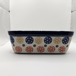 May include: A rectangular ceramic baking dish with a dark blue rim. The exterior features a repeating pattern of large beige, dark blue, and reddish-orange polka dots, interspersed with smaller dark green dots on a cream-colored background.
