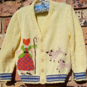 以下が含まれることがあります： 羊飼い女と羊のデザインが施された黄色のニットカーディガン。カーディガンはVネックでボタン留めです。