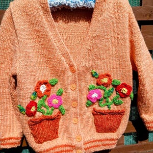 May include: A peach-colored knitted cardigan with crocheted flower embellishments. The cardigan has a V-neck, long sleeves, and a button closure. The flowers are in shades of red, pink, orange, and green.