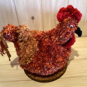 May include: A handmade, plush chicken figurine in shades of red, orange, and brown. The chicken has a red comb and wattle, a black beak, and a round, cork base. The body is made of textured yarn, giving it a fluffy appearance.