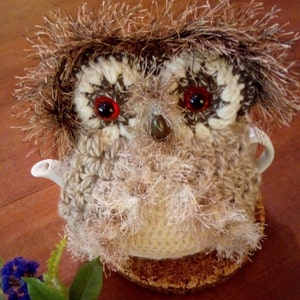 May include: A crocheted owl teapot cosy made with brown and white yarn and red eyes. The cosy is sitting on a wooden coaster.