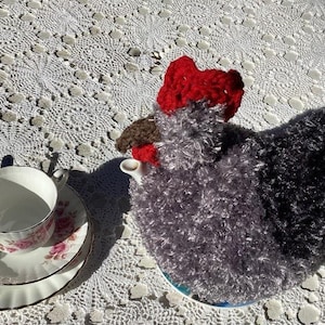 Op de afbeelding: Een handgemaakte theemuts in de vorm van een kip, met een rode kam en een grijs en zwart lichaam. Een witte kop en schotel set met roze bloemenaccenten staat ernaast op een wit kanten tafelkleed. Een lepel rust op de schotel.