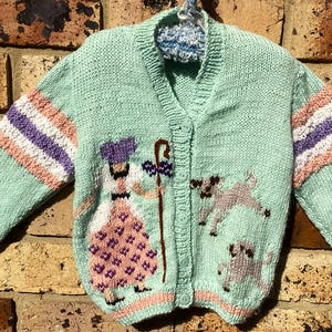 May include: A mint green knitted cardigan featuring a button-up front and long sleeves with horizontal stripes in white, purple, and pink. The front displays an intarsia design of a figure with a staff and two small dogs in grey and black.