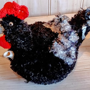 May include: A handcrafted chicken tea cosy. The cosy is mainly black with red detailing on the comb and wattle. Silver tinsel is used for the wings and tail. The beak is grey, and the eye is a small white bead.
