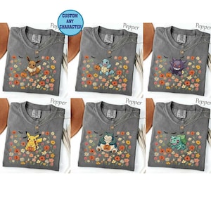 Könnte beinhalten: Sechs anthrazitfarbene T-Shirts mit Rundhalsausschnitt, die jeweils eine andere Zeichentrickfigur mit einem Halloween-Kürbis zeigen. Die T-Shirts sind mit einem bunten Blumenmuster aus orangefarbenen, gelben und rosa Blüten sowie kleinen schwarzen Fledermäusen verziert.
