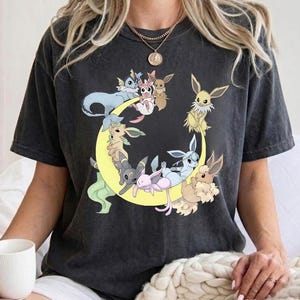 Op de afbeelding: Een antracietgrijs T-shirt met korte mouwen en een grafische print van verschillende Eevee-evolutiekarakters gerangschikt rond een grote gele halve maan. Het shirt heeft een ronde hals en een relaxte pasvorm.
