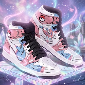 May include: High-top sneakers featuring a white, pink, and light blue color scheme with black laces. The design includes a graphic of the Sylveon character, a Poké Ball icon on the ankle collar, and decorative bow motifs on the side panels.