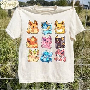 Op de afbeelding: Een ivoorkleurig t-shirt met een raster van negen kleurrijke cartoonwezens in vierkante kaders. De afbeelding bevat diverse gestileerde figuren met opvallende kenmerken zoals puntige oren, staarten en expressieve gezichten in drie rijen.