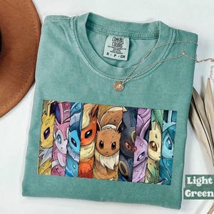 Peut inclure: Un t-shirt vert clair avec une impression graphique rectangulaire représentant neuf personnages évolutions d'Évoli stylisés disposés en panneaux verticaux. Le t-shirt a un col rond et une étiquette de marque Comfort Colors imprimée à l'intérieur.