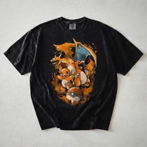 May include: A black short-sleeve t-shirt featuring a graphic print of three orange dragon-like creatures in various stages of evolution, surrounded by orange flames and a circular white and red ball icon centered on the chest area.