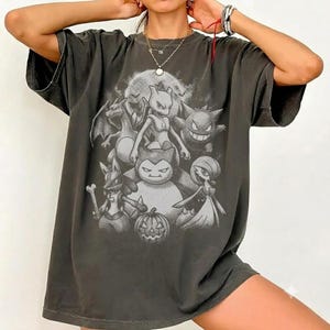 May include: A charcoal grey short-sleeved t-shirt featuring a monochromatic graphic print of various animated creatures, including a central Snorlax, Mewtwo, and a jack-o'-lantern, set against a full moon background with a distressed, vintage-style finish.