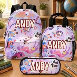 Op de afbeelding: Een bijpassende set bestaande uit een rugzak, een lunchtas en een etui, allemaal met een lichtpaarse achtergrond, een kleurrijk cartoonfiguurpatroon en de naam ANDY in dikke, donkerrode hoofdletters op elk item gedrukt.