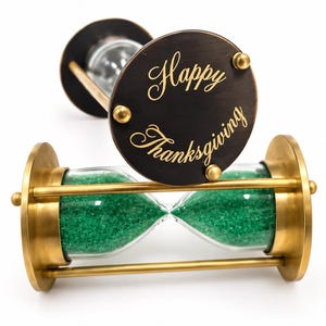 Könnte beinhalten: Zwei horizontale Sanduhren mit Messingrahmen und klaren Glaskolben, die mit grünem Sand gefüllt sind. Die vordere Sanduhr hat eine kreisförmige dunkle Metallplatte mit der goldenen Gravur Happy Thanksgiving, befestigt durch drei kleine Messingnieten.