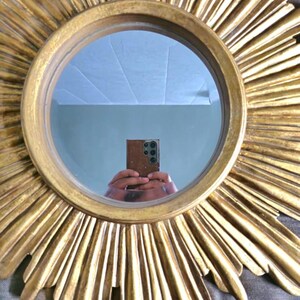 May include: A round, gold-colored mirror with a sunburst design. The mirror's frame features radiating, textured rays. The mirror reflects a room with a light-colored ceiling and a person holding a phone.