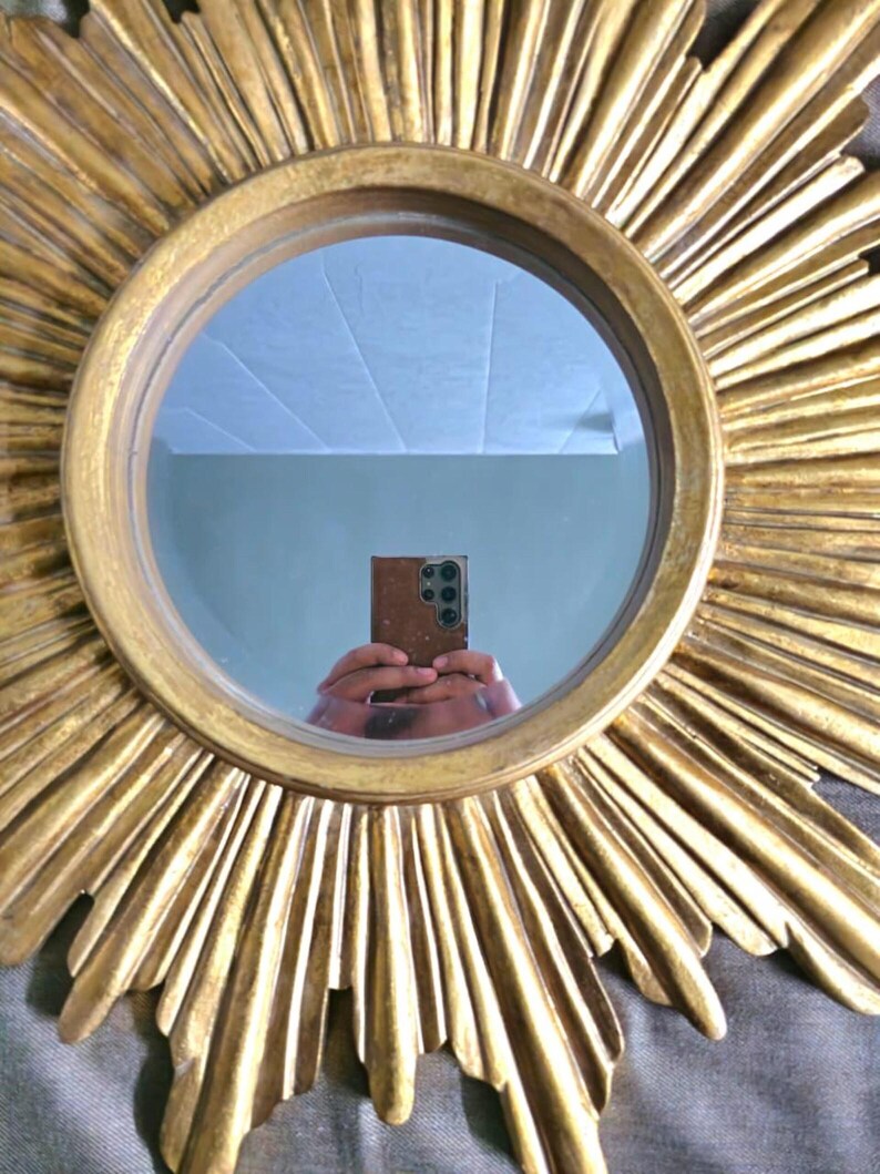 May include: A round, gold-colored mirror with a sunburst design. The mirror's frame features radiating, textured rays. The mirror reflects a room with a light-colored ceiling and a person holding a phone.