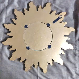 May include: Back of a gold-colored sunburst mirror. The mirror has a circular center and a decorative, irregular edge. The back features a metal hanging bracket and four blue mounting clips. The mirror is designed to be hung on a wall.