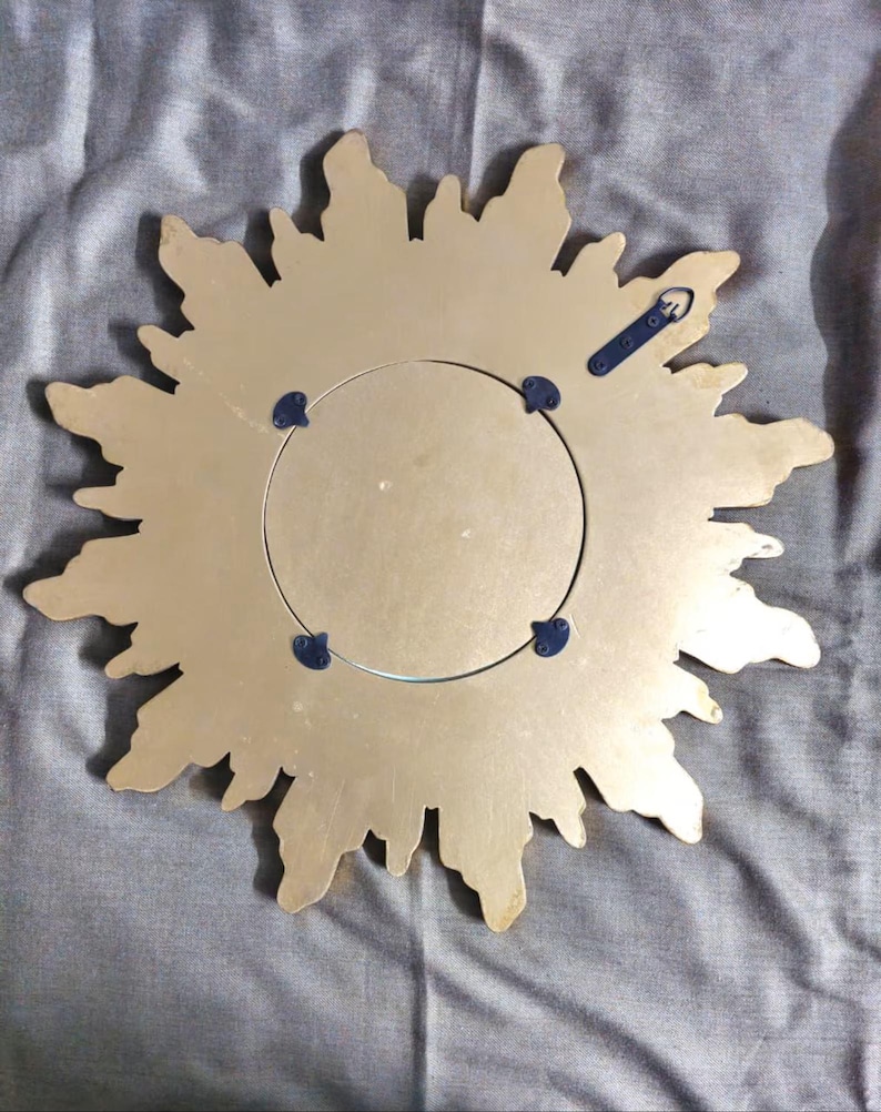 May include: Back of a gold-colored sunburst mirror. The mirror has a circular center and a decorative, irregular edge. The back features a metal hanging bracket and four blue mounting clips. The mirror is designed to be hung on a wall.