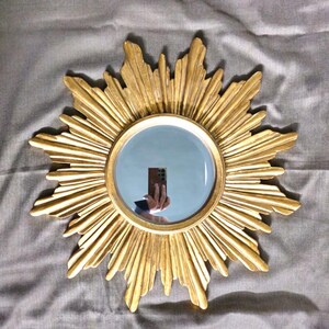 May include: A gold-colored sunburst mirror with a round, reflective center. The mirror's frame features a detailed, textured design radiating outwards. The mirror is set against a gray fabric background, creating a striking contrast.