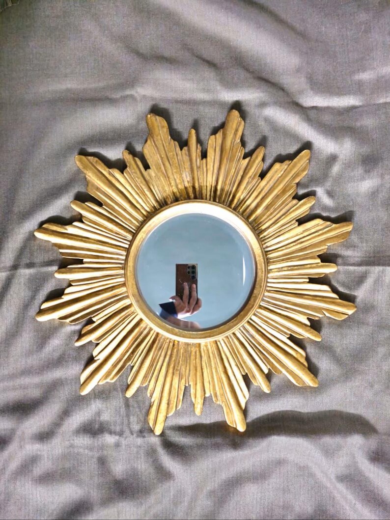 May include: A gold-colored sunburst mirror with a round, reflective center. The mirror's frame features a detailed, textured design radiating outwards. The mirror is set against a gray fabric background, creating a striking contrast.
