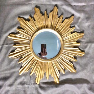 May include: A gold-colored sunburst mirror with a round, reflective center. The mirror's frame features a detailed, textured design radiating outwards. The mirror is a decorative item, suitable for wall decor.