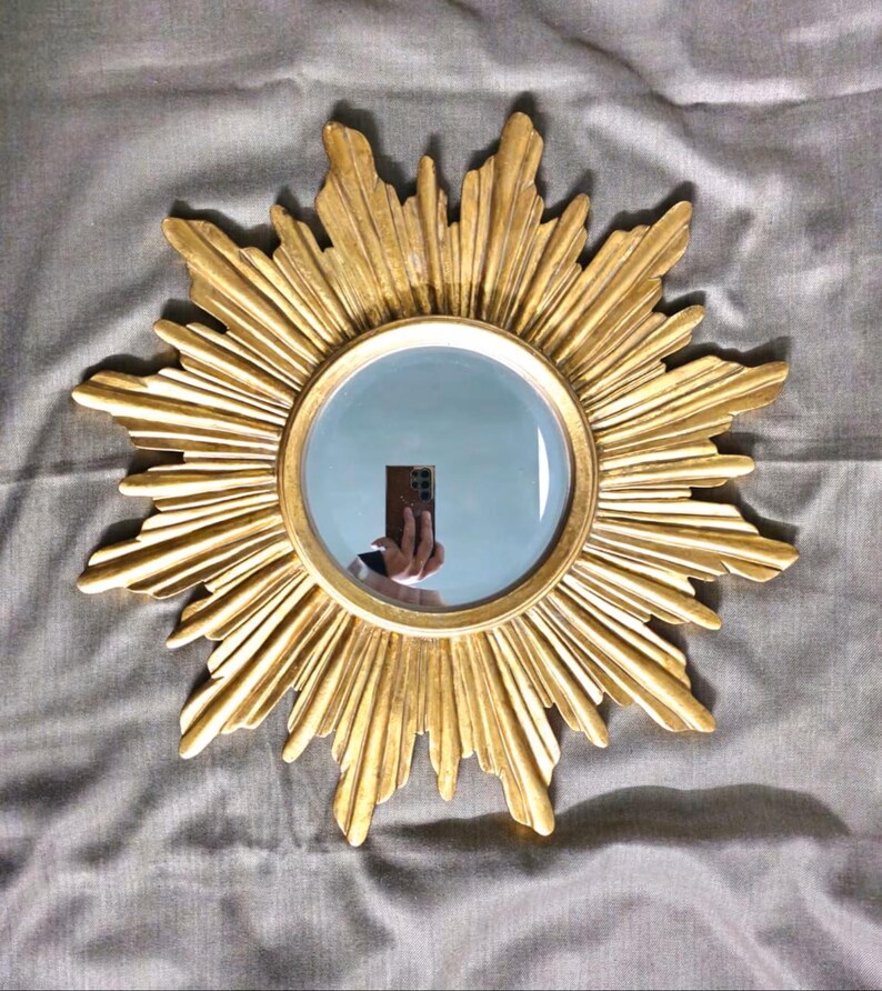 May include: A gold-colored sunburst mirror with a round, reflective center. The mirror's frame features a detailed, textured design radiating outwards. The mirror is a decorative item, suitable for wall decor.
