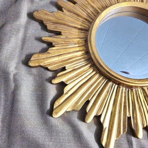 May include: A gold-colored sunburst mirror with a round, reflective center. The frame features radiating rays with a textured, aged appearance. The mirror is a decorative wall hanging.