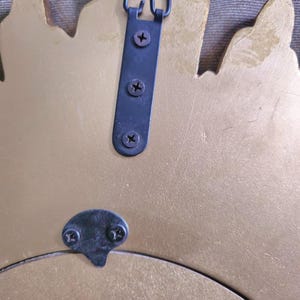 May include: Back of a gold-colored decorative mirror with a black metal hanging bracket and mounting hardware. The bracket is attached with three screws, and a smaller bracket is at the bottom.