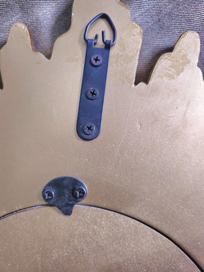 May include: Back of a gold-colored decorative mirror with a black metal hanging bracket and mounting hardware. The bracket is attached with three screws, and a smaller bracket is at the bottom.