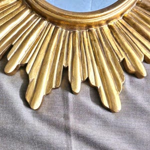 May include: Close-up of a gold-colored sunburst mirror. The mirror's frame features a detailed, textured design with radiating rays. The mirror has a circular center and is set against a neutral background.