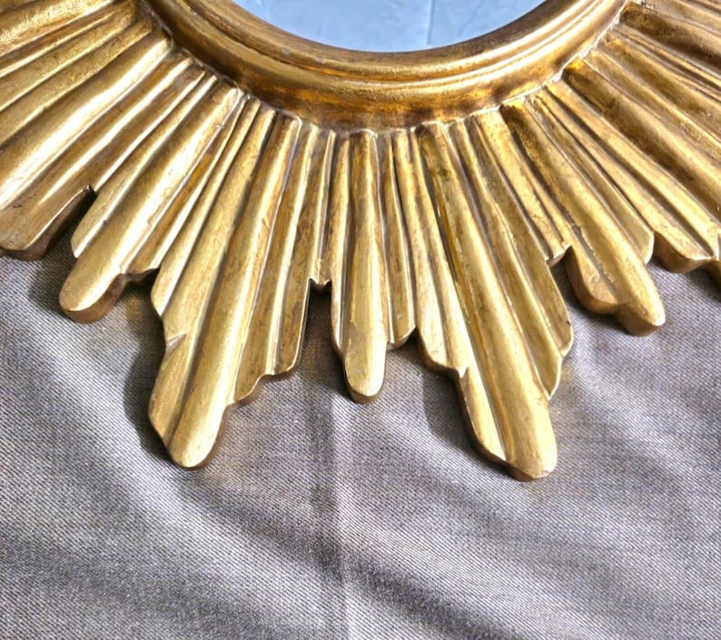 May include: Close-up of a gold-colored sunburst mirror. The mirror's frame features a detailed, textured design with radiating rays. The mirror has a circular center and is set against a neutral background.