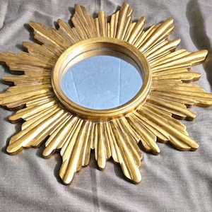 May include: A gold-colored sunburst mirror with a round, reflective center. The mirror's frame features a detailed, textured design with radiating rays. The mirror is a decorative item, suitable for wall decor.