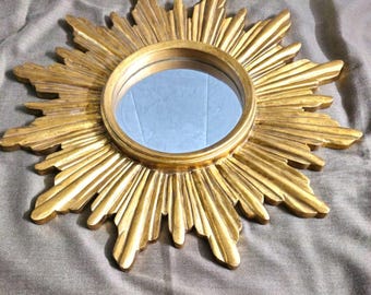 May include: A gold-colored sunburst mirror with a round, reflective center. The mirror's frame features a detailed, textured design with radiating rays. The mirror is a decorative item, suitable for wall decor.