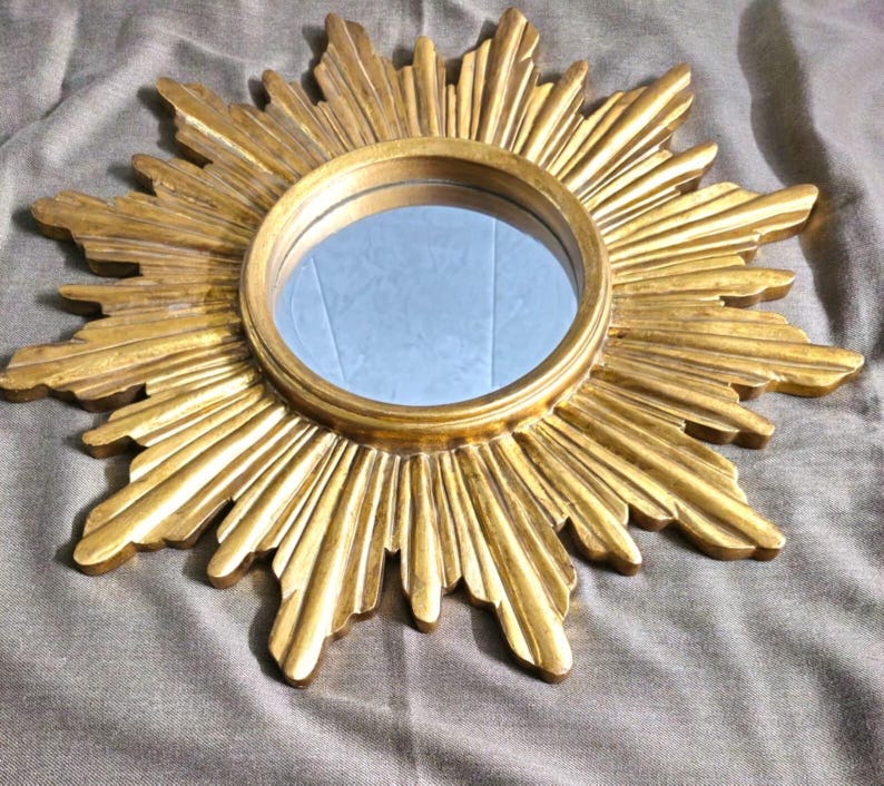 May include: A gold-colored sunburst mirror with a round, reflective center. The mirror's frame features a detailed, textured design with radiating rays. The mirror is a decorative item, suitable for wall decor.