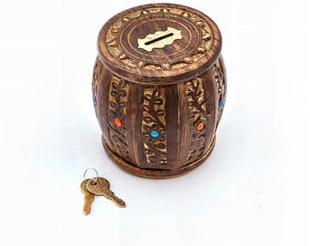 Puede incluir: Un banco de dinero de madera en forma de barril con intrincadas tallas florales y piedras preciosas incrustadas. El banco presenta una ranura para insertar monedas y una pequeña placa de latón. Dos llaves se colocan junto al banco. El banco es marrón con detalles dorados.