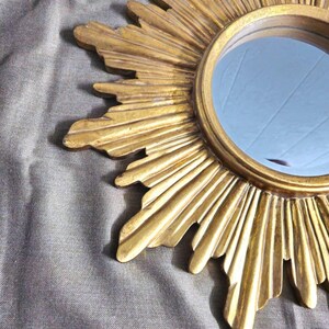 May include: A gold-colored sunburst mirror with a round, reflective center. The frame features radiating rays with textured details. The mirror is a decorative accent piece.
