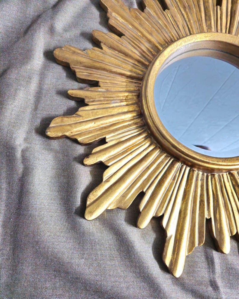 May include: A gold-colored sunburst mirror with a round, reflective center. The frame features radiating rays with textured details. The mirror is a decorative accent piece.