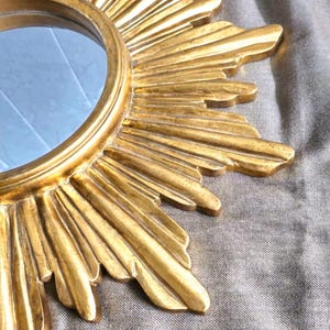 May include: A gold-colored sunburst mirror with a round central mirror. The frame has a detailed, textured design with radiating rays. The mirror is a decorative wall hanging.