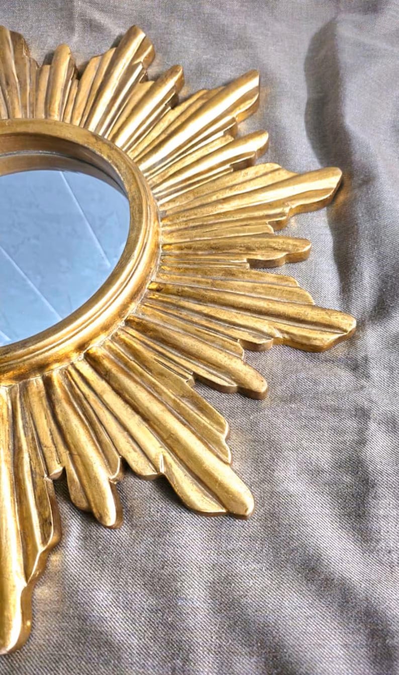May include: A gold-colored sunburst mirror with a round central mirror. The frame has a detailed, textured design with radiating rays. The mirror is a decorative wall hanging.