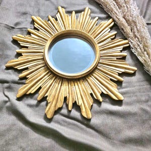 May include: A gold-colored sunburst mirror with a round mirror in the center. The sunburst design features radiating rays. The mirror is a decorative wall hanging.