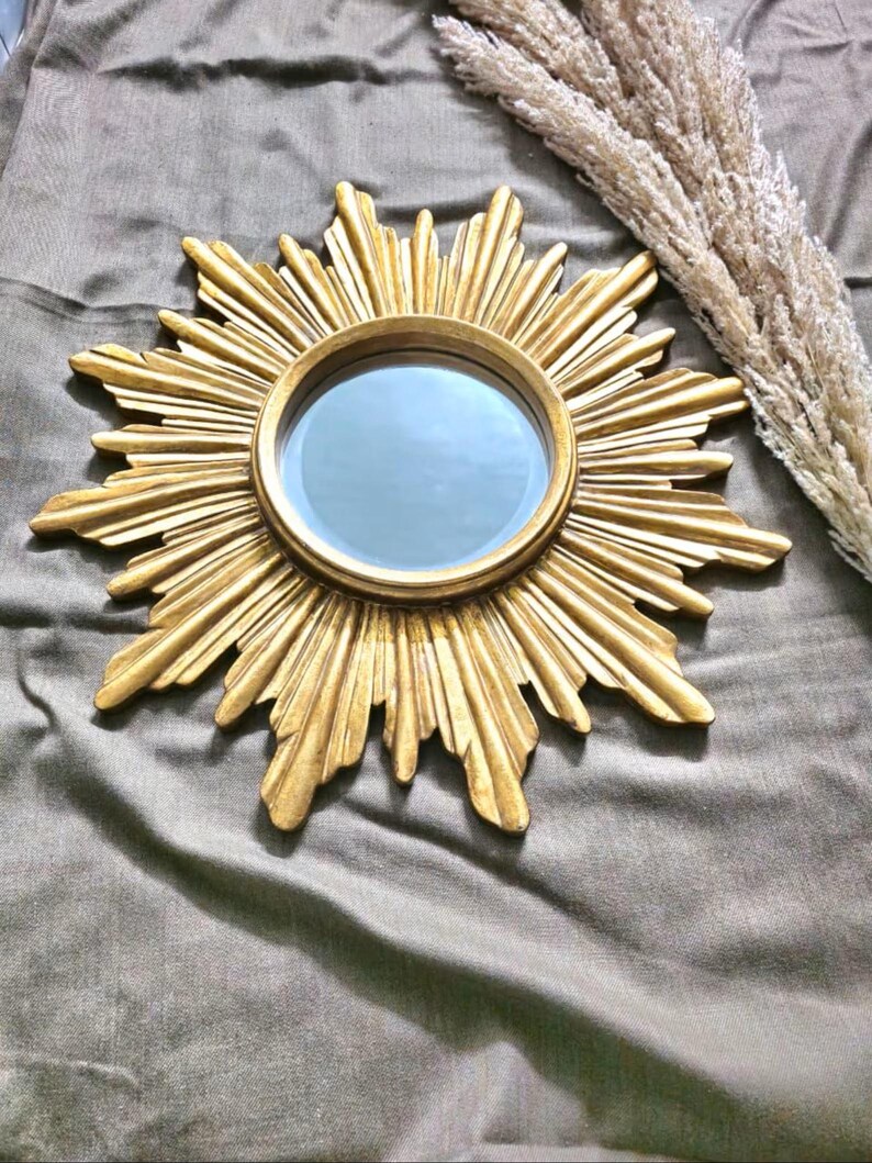 May include: A gold-colored sunburst mirror with a round mirror in the center. The sunburst design features radiating rays. The mirror is a decorative wall hanging.