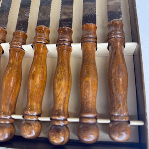 May include: A set of six steak knives with serrated stainless steel blades and brown wooden handles. Each blade is stamped with the text Stainless Steel Japan. The knives are arranged in a row within a rectangular cardboard storage box.