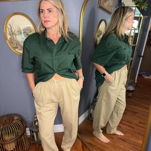 May include: A dark green short-sleeved button-down shirt paired with khaki-colored drawstring pants. The shirt has a chest pocket. The pants have pockets and a drawstring waist. The outfit is casual and comfortable.