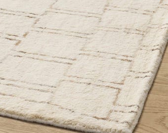May include: A plush area rug featuring a cream-colored base with a repeating geometric pattern of thin, light brown rectangular outlines. The rug has a soft, textured pile surface that covers the floor, displaying a consistent grid-like design.