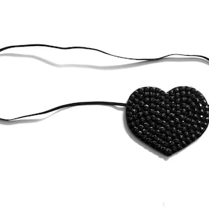 May include: A black heart-shaped eye patch with a black elastic strap. The heart is densely covered in small, dark, reflective elements. The eye patch is designed to be worn over one eye.