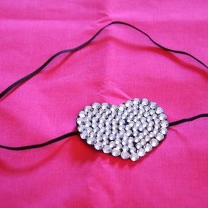 Könnte beinhalten: Ein schwarzes Stirnband mit herzförmigem Schmuck. Das Herz ist mit vielen kleinen, klaren Strasssteinen besetzt. Das Stirnband liegt auf einem leuchtend pinkfarbenen Hintergrund.