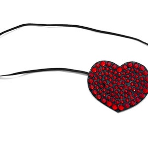 May include: A heart-shaped eye patch with a black elastic band. The patch is covered in numerous red rhinestones. The design is set against a white background.