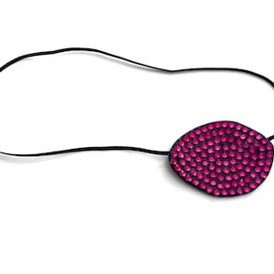 Könnte beinhalten: Eine dekorative Augenklappe mit einem schwarzen Gummiband. Die Augenklappe hat eine unregelmäßige ovale Form und ist mit kleinen, leuchtend rosa Strasssteinen bedeckt. Der Hintergrund ist schlicht weiß.