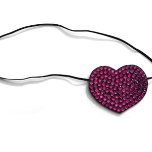 Puede incluir: Un parche para el ojo en forma de corazón con una banda elástica negra. El parche está cubierto de pequeños pedrería rosa brillante. El parche para el ojo está sobre un fondo blanco.